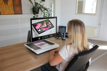 Back view of a woman operating the Acuity Speech desktop magnifier, showing the adjustable arm and widescreen display.