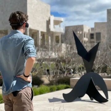 A man in a blue shirt, wearing an ARx Vision headset, is observing a black abstract sculpture. He is facing away from us.