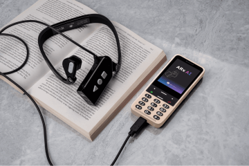 The image shows the ARx Vision headset resting on an open book. Next to it is a Golden Sand BlindShell Classic 3 phone connected to the headset via a cable. The phone screen displays the ARx AI app interface. The headset is positioned with its tactile buttons visible, and the book is open, with the device laid across the pages. The scene is set on a gray surface.
