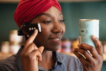 A woman wearing a red headscarf is using the ARx Vision device attached to her temple. She is holding a can in her hand and appears to be reading or examining it. The ARx Vision device, with tactile buttons, is positioned near her cheek, secured by a headband. She is smiling and focused on the item she is observing.