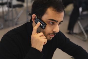 A close-up image of a man wearing an ARx Vision headset, a bone-conducting device held to his ear. The headset transforms a smartphone into a live-streaming camera. The man is focused while using the device, with a blurred indoor background.