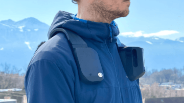 This image shows a person wearing a dark blue jacket with a high collar, outdoors with a snowy mountain landscape in the background. On the person's shoulders, there are two black, rectangular devices attached, one on each side. These devices seem to be technological gadgets, possibly for capturing video or audio, or perhaps wearable air purifiers or cooling devices. The person appears to be standing in a cold environment, indicated by the snowy mountains and the winter jacket.