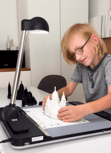 A young boy with glasses is interacting with a 3D model of the Sacré-Cœur Cathedral in Paris. The model is placed on a tactile graphic displayed on the Tactonom Reader. A desk lamp illuminates the workspace. In the background, more 3D models of different buildings are visible, arranged on a desk. The child is focused, exploring the tactile model with his hands.