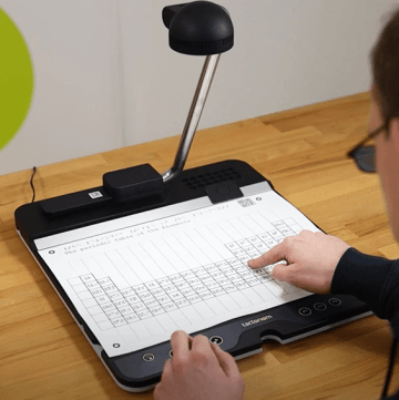 A person using Tactonom Reader with a periodic table of elements displayed on a tactile surface. Tactonom has a built-in camera and tactile buttons for navigation. The individual is pointing at the periodic table while seated at a wooden desk.