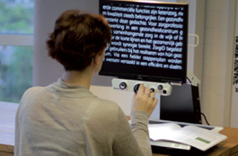 A woman using the iSee desktop magnifier at a table, adjusting the front control knobs while enlarged white text on a black background is displayed on the screen.