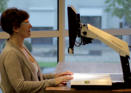 A woman sitting at a desk using the iSee desktop magnifier, shown from the side with the adjustable arm and screen positioned above the reading platform.